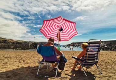 Tenerife Adası, İspanya - 6 Kasım 2015: Yaşlı erkek ve kadın şemsiyesi altında adada Tenerife, Kanarya Adaları San Juan plajda güneşlenirken