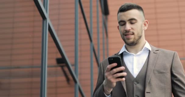 Ο άνθρωπος χρησιμοποιώντας επιχειρηματική εφαρμογή στο smart phone περπατώντας στην πόλη. Όμορφος νεαρός επιχειρηματίας επικοινωνία για smartphone χαμογελώντας αυτοπεποίθηση — Αρχείο Βίντεο