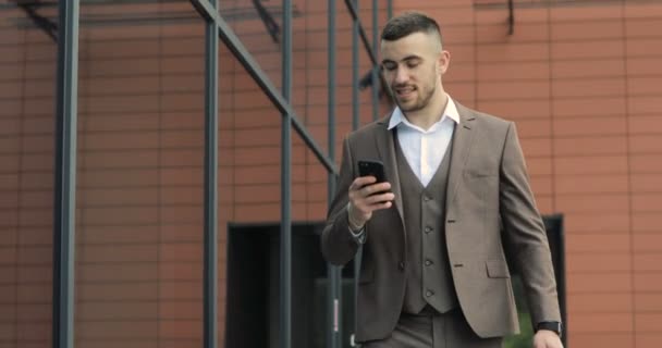 Чоловік використовує бізнес-додаток на смартфоні, що ходить по місту. Красивий молодий бізнесмен спілкується на смартфоні посміхаючись впевнено — стокове відео