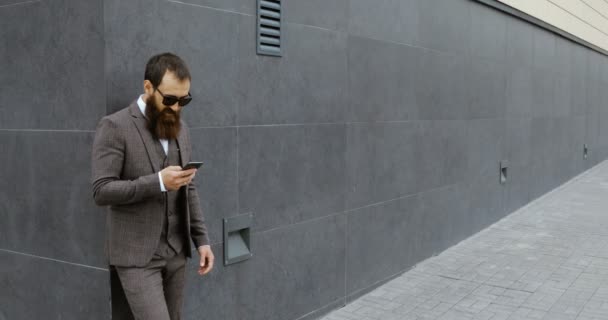 Atractivo hombre de negocios barbudo mirando a su alrededor y utilizando su teléfono inteligente mientras se mantiene fuera del edificio moderno , — Vídeo de stock