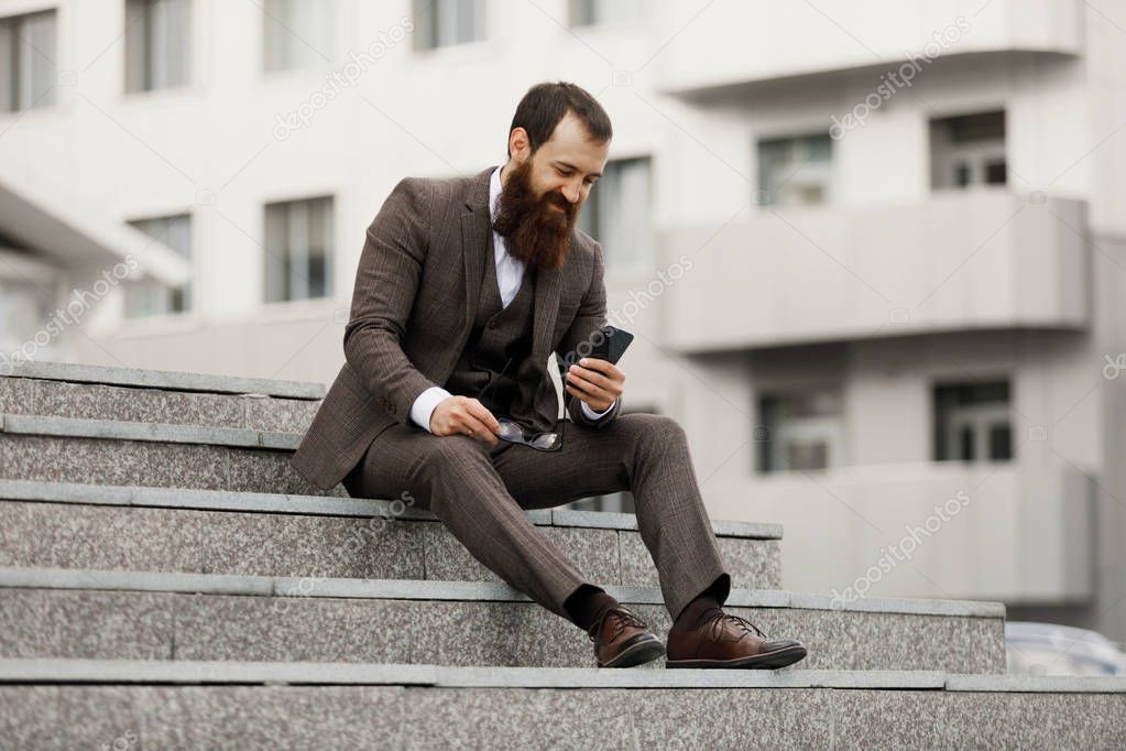 Businessman using mobile phone app texting outside of office in urban city with skyscrapers buildings in the background. Young caucasian man holding smartphone for business work.