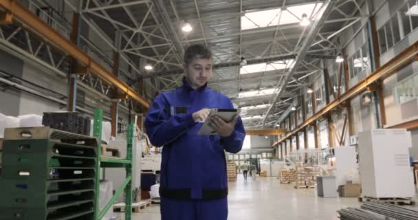 Un uomo sta lavorando in una fabbrica con un tablet digitale, è vestito con un'uniforme blu. operaio di fabbrica . — Video Stock