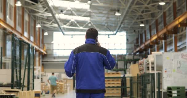 Ingeniero en uniforme azul está caminando a través de fábrica y hablando en el teléfono móvil. Vista trasera — Vídeo de stock