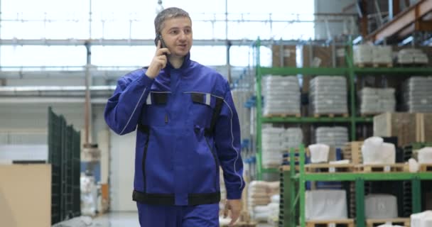 Ingeniero en uniforme azul está caminando a través de fábrica y hablando en el teléfono móvil. Vista frontal — Vídeos de Stock