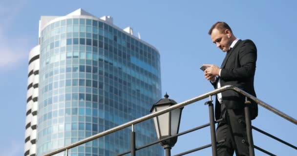 Смс-повідомлення людини, використовуючи додаток на смартфоні в місті. Красивий молодий бізнесмен використовує смартфон, посміхаючись щасливим. Міська чоловіча професійна комунікація в його 30-х роках проти хмарочоса і неба — стокове відео