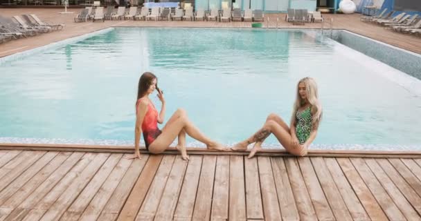 Dos chicas posando junto a la piscina del hotel. Mujeres jóvenes en un traje de baño de una sola pieza sentadas en el borde de una piscina, posando y sonriendo . — Vídeo de stock