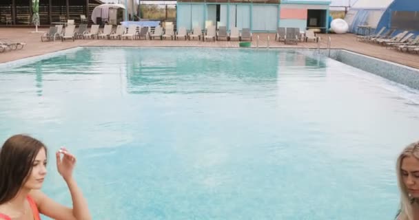 Dos chicas posando junto a la piscina del hotel. Mujeres jóvenes en un traje de baño de una sola pieza sentadas en el borde de una piscina, posando y sonriendo . — Vídeo de stock