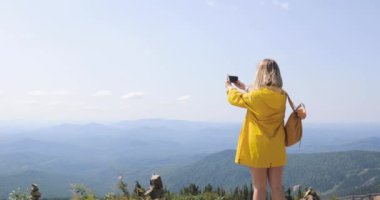 Kız sarı yağmurluk içinde bir fotoğraf bir dağ, mavi gökyüzü ve dağlar bir arka plan üzerine bir smartphone ayakta yapar