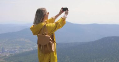 Kız sarı yağmurluk içinde bir fotoğraf bir dağ, mavi gökyüzü ve dağlar bir arka plan üzerine bir smartphone ayakta yapar
