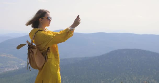 Дівчина в жовтому плащі робить фотографію на смартфоні, що стоїть на вершині гори, блакитного неба та гір на фоні — стокове відео