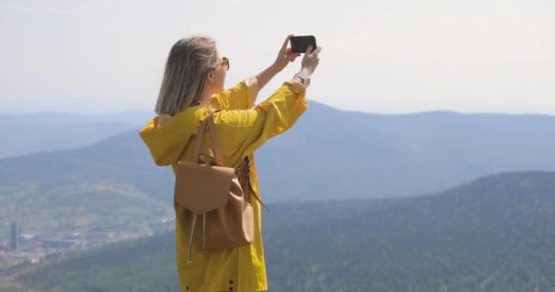Flicka i en gul regnrock gör ett foto på en smartphone stående ovanpå ett berg, blå himmel och berg på en bakgrund — Stockvideo