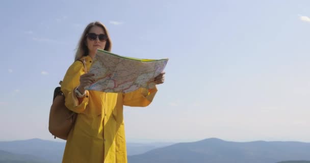 Eine junge Frau wandert in Regenjacke auf einem Bergpfad, hält an und prüft die Wegbeschreibung. Wanderer erreicht Berggipfel, prüft Wegbeschreibung auf Karte — Stockvideo