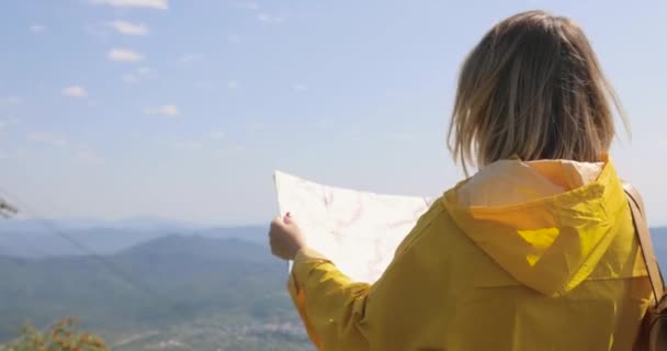 Visão traseira do caminhante feminino caucasiano em capa de chuva amarela fica nas montanhas com um mapa na mão — Vídeo de Stock