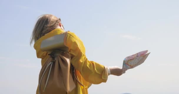 Vista posterior de la mujer senderista caucásica en impermeable amarillo se encuentra en las montañas con un mapa en la mano — Vídeos de Stock