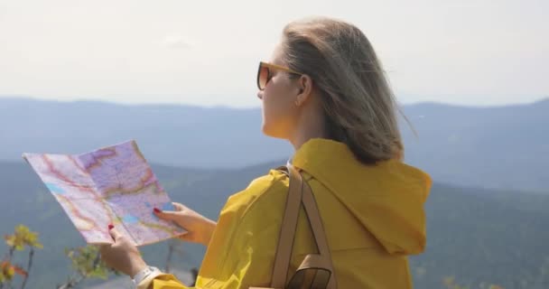 Caminhante em uma capa de chuva amarela olhando para o mapa do topo da montanha. mulher com mapa em montanhas 4k — Vídeo de Stock