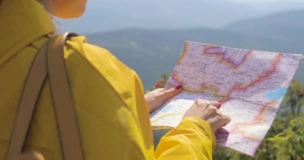 Vista posterior de la mujer senderista caucásica en impermeable amarillo se encuentra en las montañas con un mapa en la mano — Vídeos de Stock