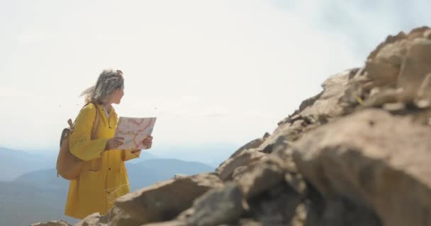 Mujer joven senderismo im impermeable amarillo con una mochila en las montañas sosteniendo mapa de papel en las manos . — Vídeo de stock