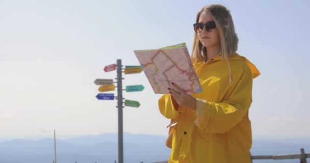 Mujer joven senderismo im impermeable amarillo con una mochila en las montañas sosteniendo mapa de papel en las manos . — Vídeos de Stock