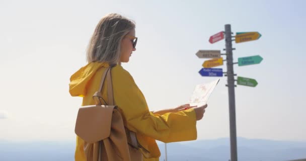 Mujer joven senderismo im impermeable amarillo con una mochila en las montañas sosteniendo mapa de papel en las manos . — Vídeos de Stock