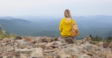 Kız sarı yağmurluk içinde güzel bir manzarayı bir dağın tepesinde oturuyor