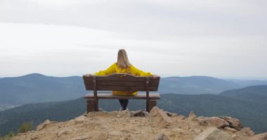 Kız sarı yağmurluk içinde güzel bir manzarayı bir dağın tepesinde oturuyor