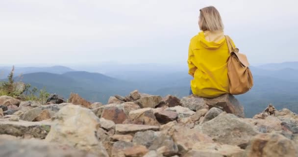 Niña en un impermeable amarillo se sienta en la cima de una montaña, disfrutando de una hermosa vista — Vídeos de Stock