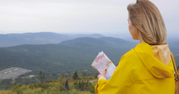 Jonge vrouwelijke wandelen im gele regenjas met een rugzak in bergen bedrijf papieren kaart in handen. — Stockvideo