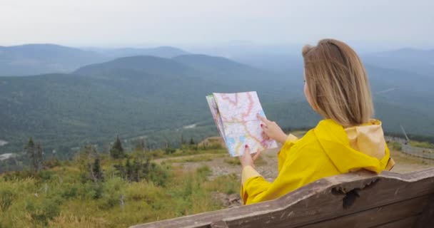 Mujer joven senderismo im impermeable amarillo con una mochila en las montañas sosteniendo mapa de papel en las manos . — Vídeos de Stock