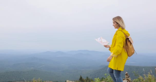 Mujer joven senderismo im impermeable amarillo con una mochila en las montañas sosteniendo mapa de papel en las manos . — Vídeos de Stock