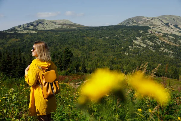 Bokovky mladá dívka v žlutá pláštěnka s batohem na vrchol hory. Turistické cestovatel na pozadí — Stock fotografie