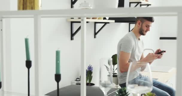 Caucásico hombre permaneciendo en la cocina y la navegación en el teléfono móvil en casa . — Vídeos de Stock