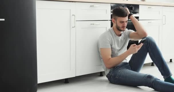 Caucásico hombre permaneciendo en la cocina y la navegación en el teléfono móvil en casa . — Vídeos de Stock