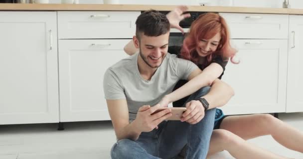 Atractiva pareja joven con teléfono inteligente en casa en la cocina — Vídeo de stock