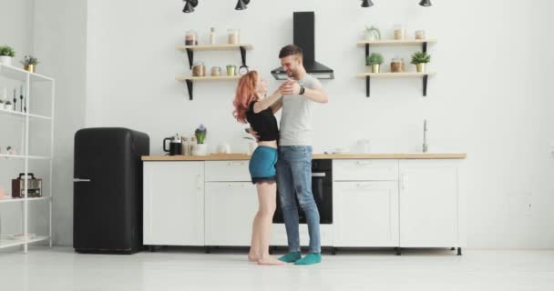 Mañana en casa feliz pareja joven recién casada bailando escuchando música en la cocina vistiendo pijamas enamorados divirtiéndose — Vídeos de Stock