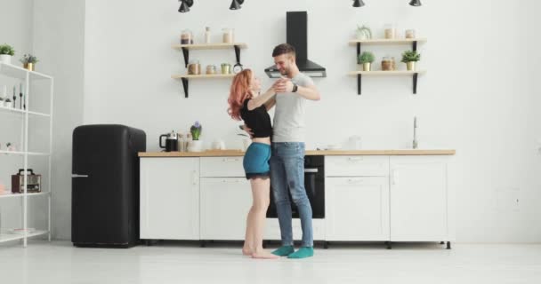 Mañana en casa feliz pareja joven recién casada bailando escuchando música en la cocina vistiendo pijamas enamorados divirtiéndose — Vídeo de stock