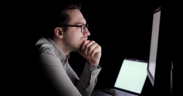 El hombre trabaja en la computadora, lee atentamente documentos en su lugar de trabajo en la oscuridad . — Vídeo de stock