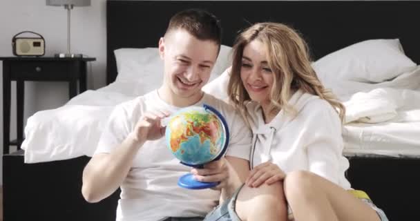 Gelukkige geliefde paar jongen en meisje zitten naast het bed zijn op zoek naar Globe kiezen van een plek om te reizen op huwelijksreis. — Stockvideo