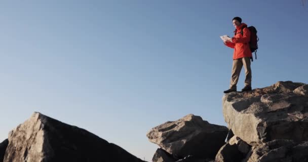 Man hiker is looking at the map in mountains checking the route. Standing on big rocks at blue sky background. — Stock Video