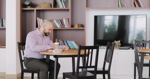 L'uomo pelato barbuto in abiti da ufficio sta leggendo un libro seduto nel bar dell'hotel con tazza di cappuccino. . — Video Stock