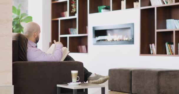 Man is reading a book in hotel hall cafe with capuccino cup sitting on the couch in front of the fireplace. — Stock Video