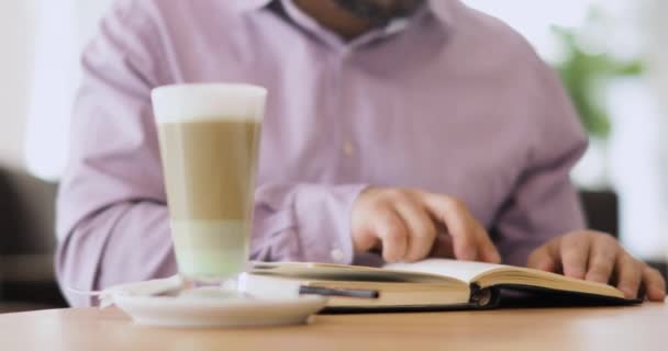 Man is reading a book with his finger on the page sitting in cafe with cup of cappuccino, front view. — Stock Video