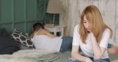 Young woman worried by problems in the relationship sitting in bed, her boyfriend is lying near turning away.