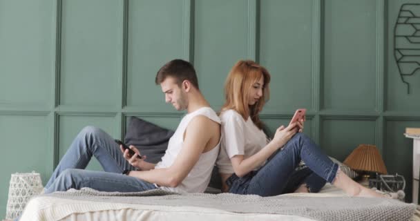 Mężczyzna i kobieta siedzą na plecach nawzajem w łóżku i przeglądając swoje telefony komórkowe. — Wideo stockowe