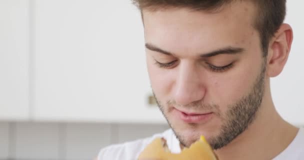 Young man is eating burger at home and talking with somebody. — Stockvideo