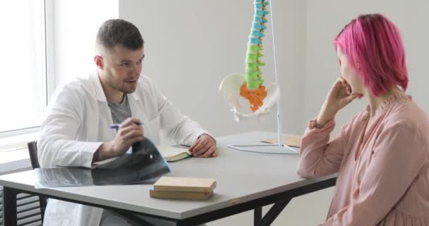 El médico está hablando con una mujer paciente con pelos rosados sobre sus problemas con la columna vertebral que muestra el área afectada en el modelo de columna vertebral . — Vídeo de stock
