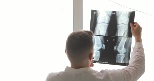 Roentgenologist está aprendiendo huesos en imágenes de rayos X de pie cerca de la ventana, vista posterior . — Vídeo de stock