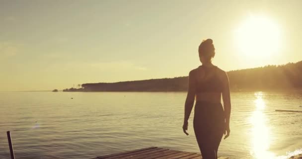 Jonge vrouw begint yoga te doen op de pier aan de rivier bij zonsondergang, achteraanzicht. — Stockvideo