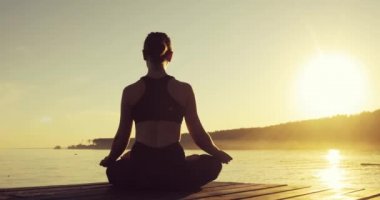 Genç kadın gün batımında denize yakın iskelede oturan Lotus Pose meditasyon.