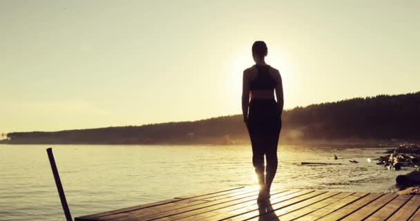 Menina desportiva está praticando ioga em pose de árvore em pé no cais pelo rio ao pôr do sol . — Vídeo de Stock