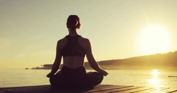 Junge Frau meditiert in Lotus-Pose und sitzt bei Sonnenuntergang auf einem Steg am Meer. — Stockvideo